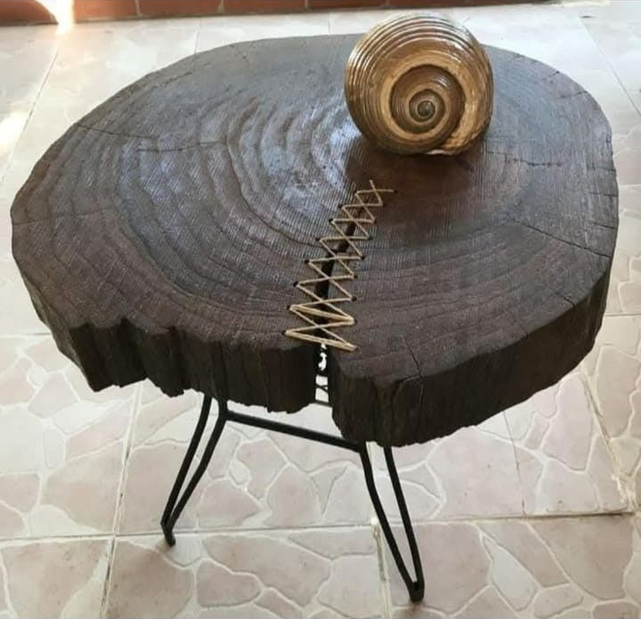 Una mesita auxiliar redonda de madera oscura con patas metálicas en forma de horquilla, que presenta una reparación decorativa con costuras y una concha en espiral.
