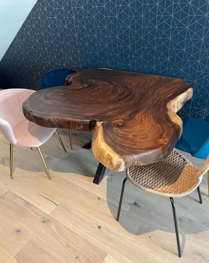 Una mesa de madera maciza con borde natural se encuentra en una habitación con papel pintado estampado, rodeada de sillas en tonos rosa, azul y marrón texturizado.