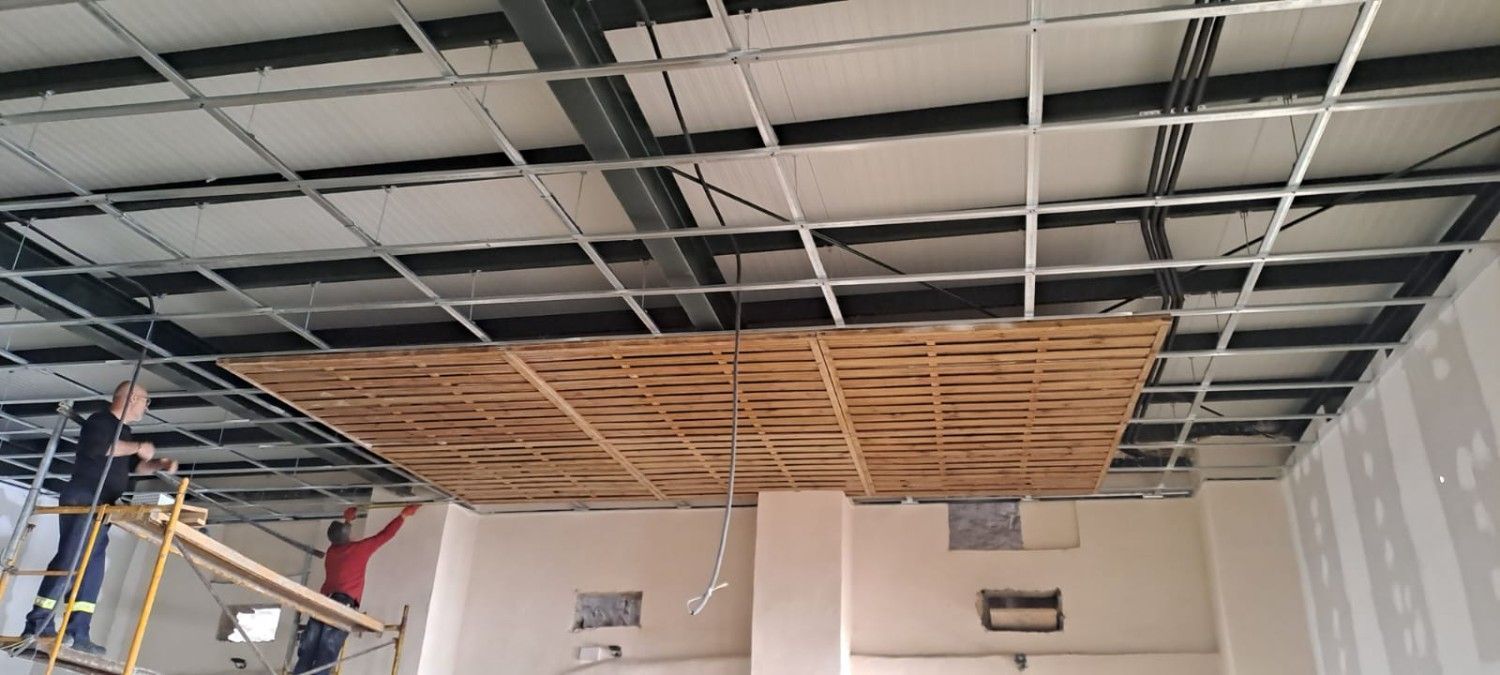 Dos obreros, subidos a un andamio, instalan paneles de madera en el techo de una estructura metálica en una habitación en construcción.