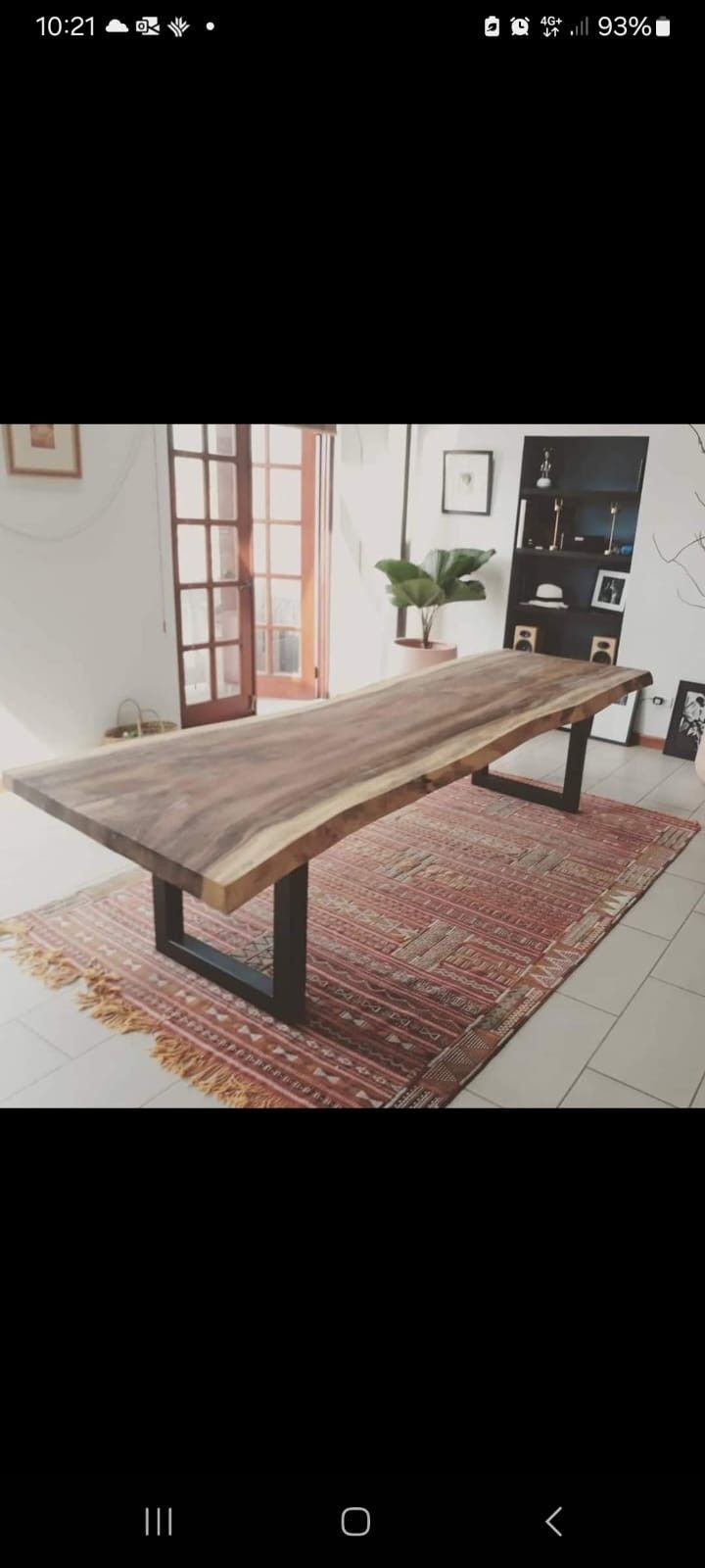 Una mesa de comedor larga de madera con bordes naturales y patas de metal oscuro en forma de U, colocada sobre una alfombra estampada en una habitación luminosa.