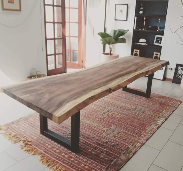 En un salón moderno, una mesa de comedor de madera con bordes naturales y patas de metal negro reposa sobre una alfombra estampada.