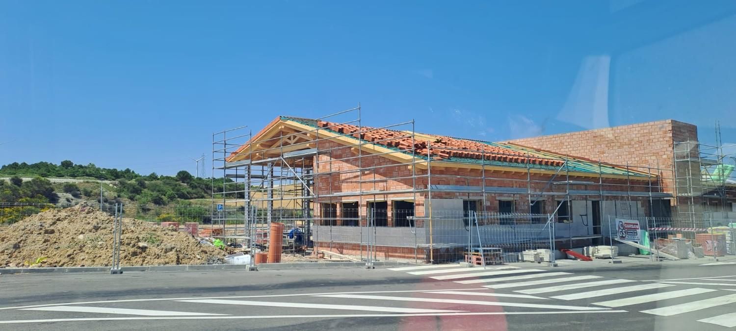 Una obra en construcción donde se observa un edificio de una sola planta parcialmente construido, con estructura de techo de madera, bajo un cielo azul despejado.