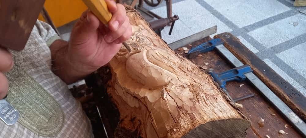 Una persona utiliza un cincel y un mazo para tallar la figura de un búho en madera, en un tronco tosco sujeto con una abrazadera en un banco de trabajo.