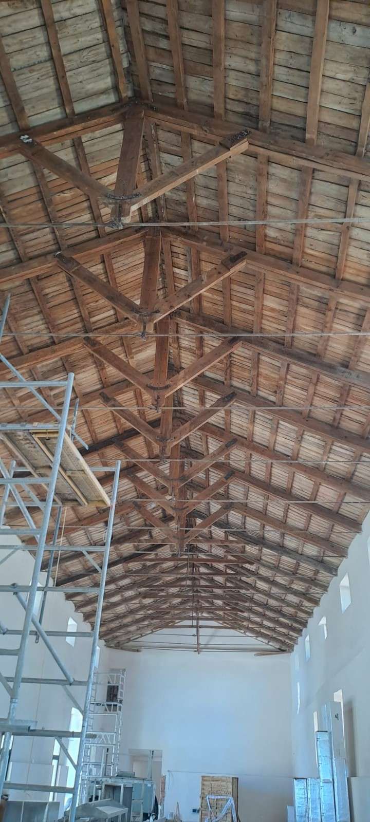 Vista interior de una habitación en construcción, que muestra un techo alto con vigas de madera sobre paredes blancas y andamios.