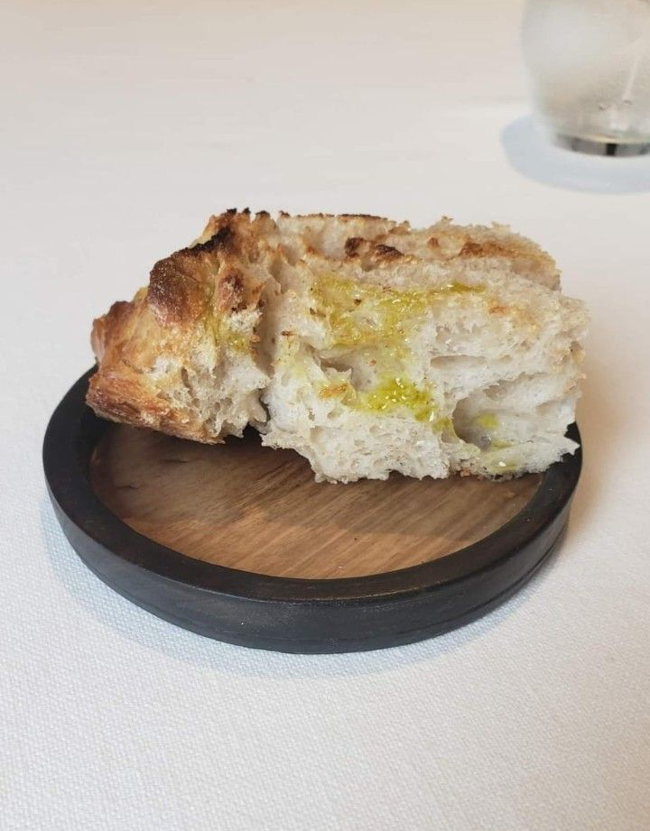 Una sola rebanada de pan crujiente rociada con aceite de oliva, servida en un pequeño plato de madera con borde oscuro.