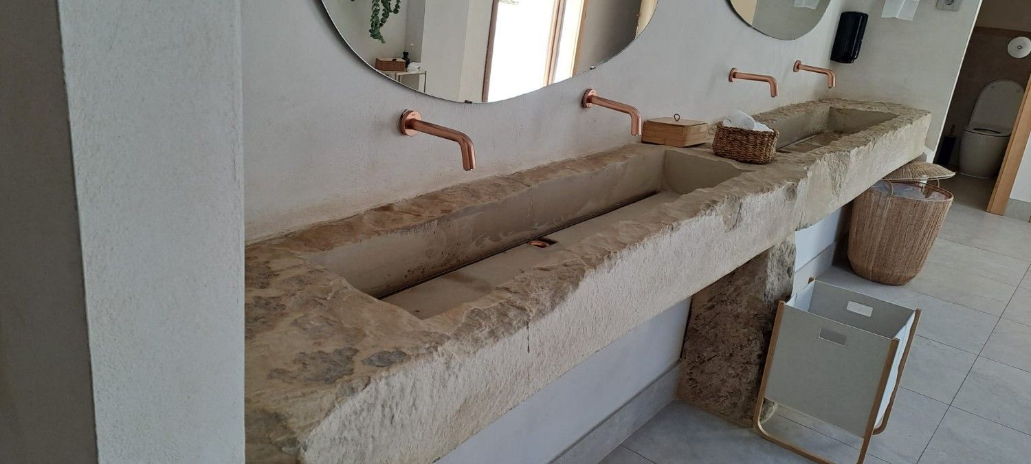 Un lavabo largo y rústico de piedra con dos grifos de cobre montados encima en un baño luminoso y moderno.