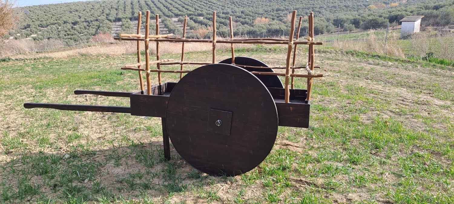 Un carro de madera con grandes ruedas macizas y una caja de listones descansa en un campo de hierba, con un huerto en la ladera de una colina al fondo.