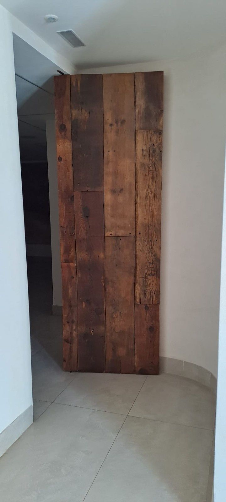 Una puerta vertical hecha de tablones de madera reciclada y teñida se alza en un pasillo con paredes blancas y un suelo de baldosas claras.