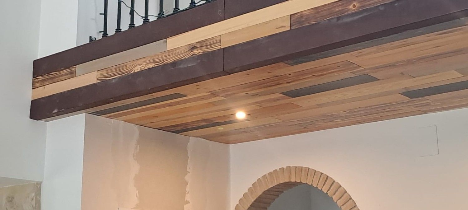Un techo con vigas de madera y tablones horizontales, una luz empotrada y un arco de ladrillo contra una pared blanca.
