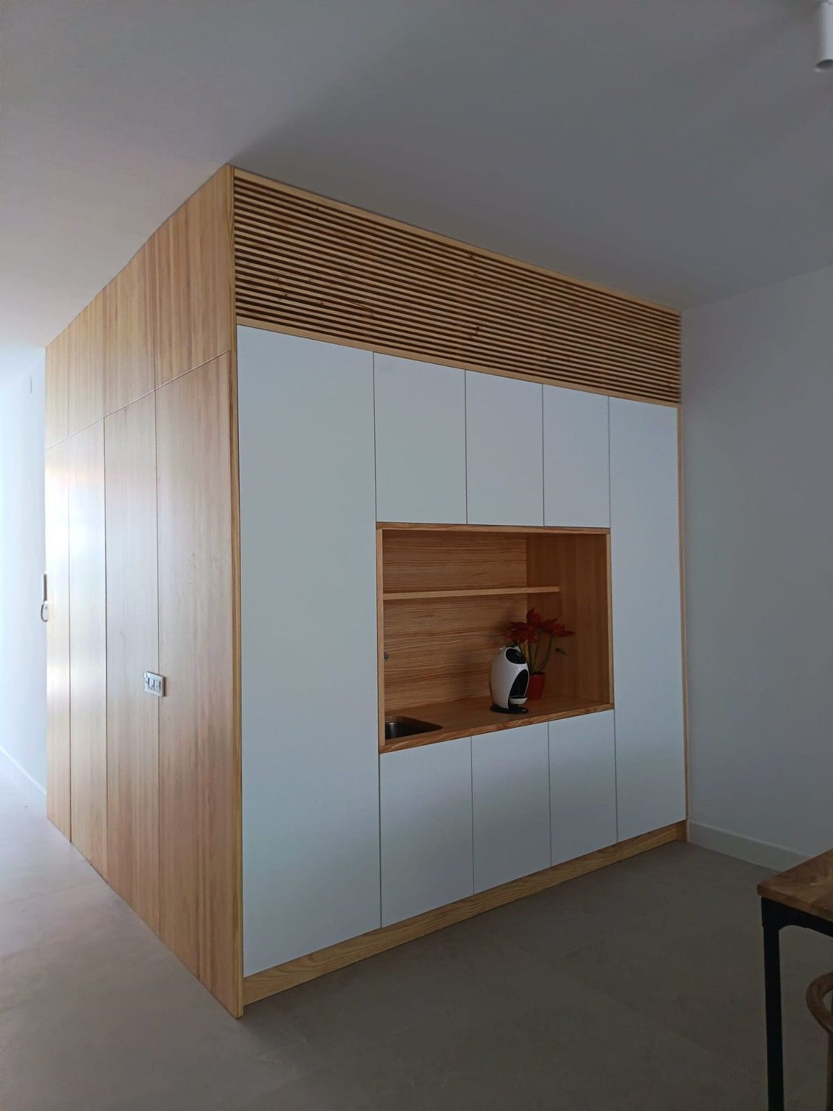 Un mueble de almacenamiento alto y modular, de madera y color blanco, con un nicho central de estanterías abiertas y un panel superior de listones, ideal para una habitación.