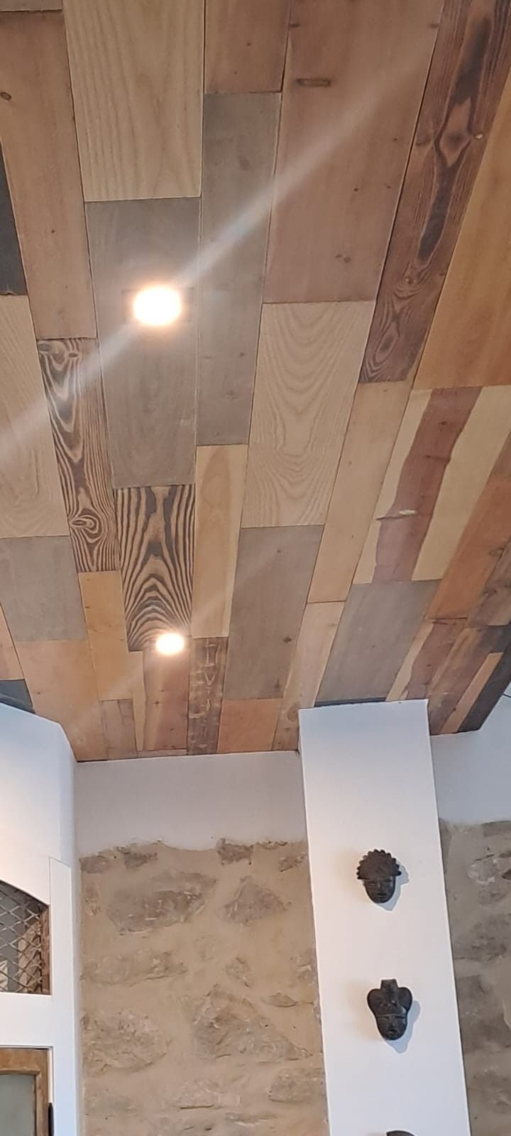 Un techo con tablones de madera desiguales y dos luces empotradas sobre una pared con mampostería de piedra y decoración oscura.
