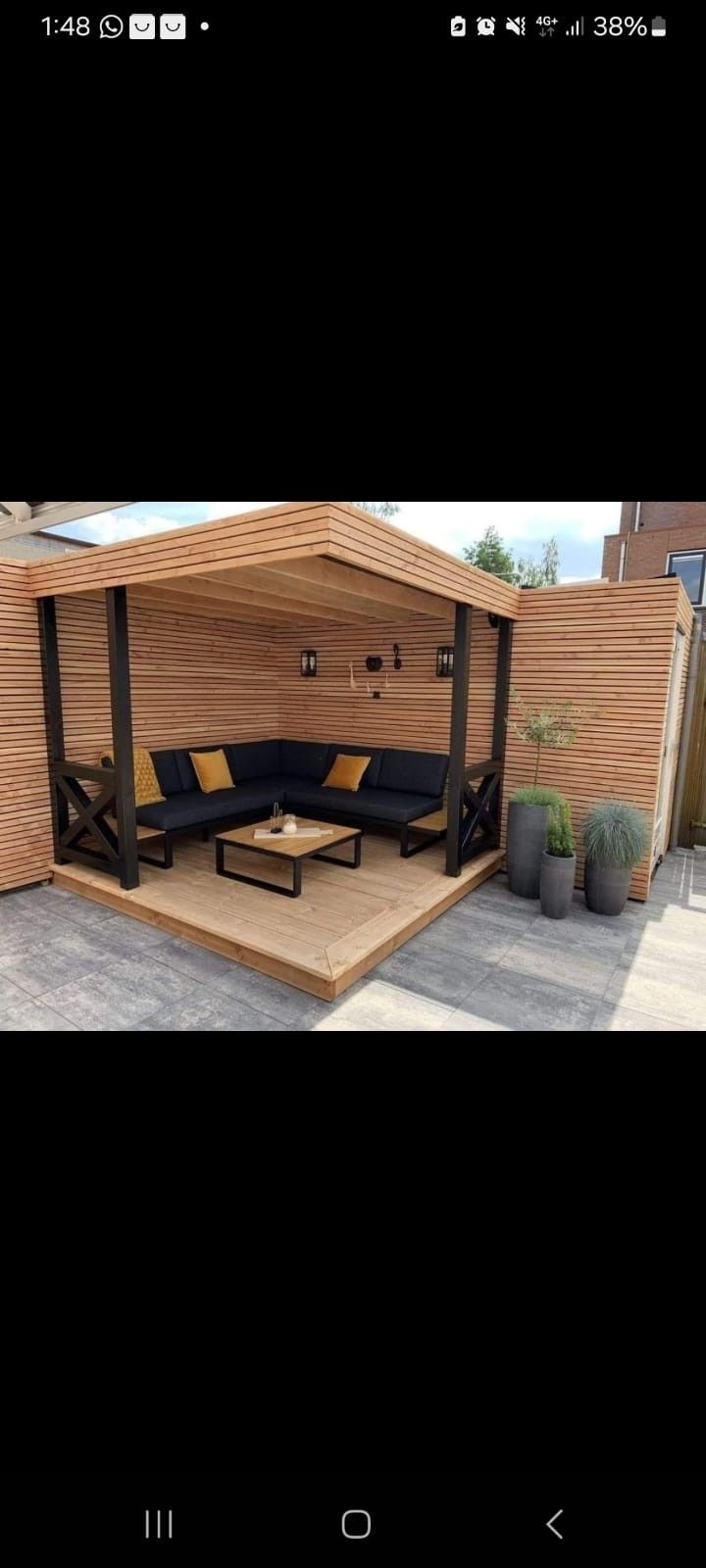 Una pérgola de madera al aire libre con un sofá esquinero negro y una mesa de centro, situada frente a muros de madera con textura ondulada que proporcionan privacidad.