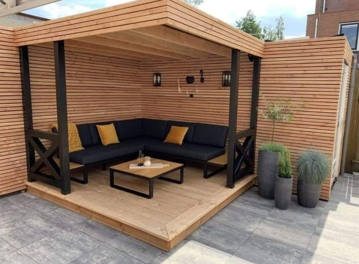 Una pérgola de madera al aire libre con un sofá esquinero negro, una mesa de centro e iluminación decorativa en un patio embaldosado.