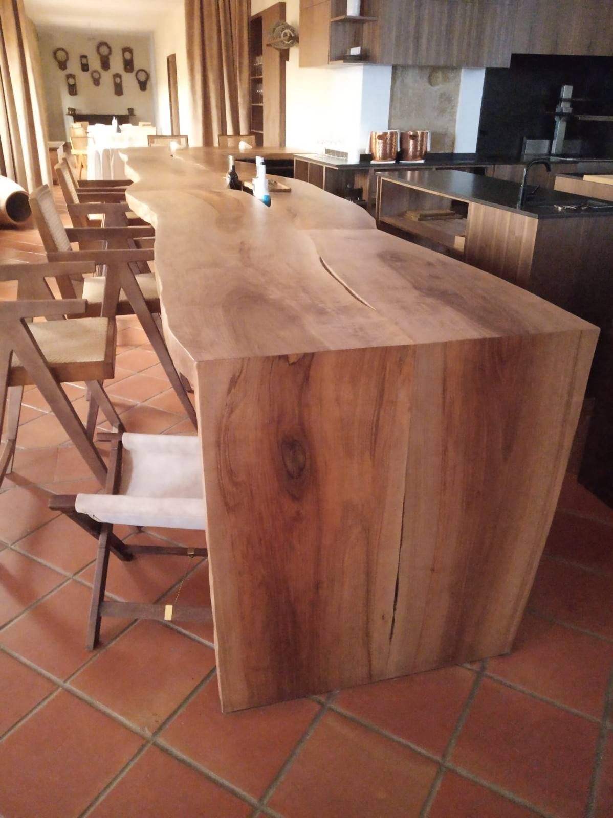 Una larga barra de madera con bordes naturales se ubica en una habitación luminosa con suelo de baldosas de terracota y sillas de madera.