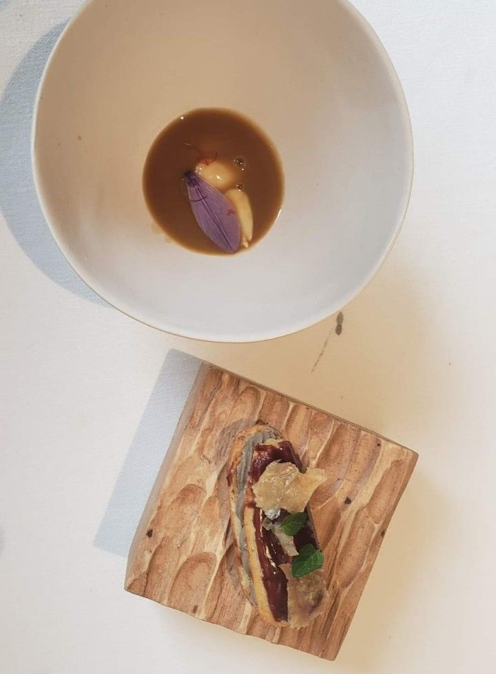 Un cuenco blanco con caldo y un pétalo de flor junto a una pequeña tabla de madera coronada con una rebanada de pan tostado adornada con hierbas.