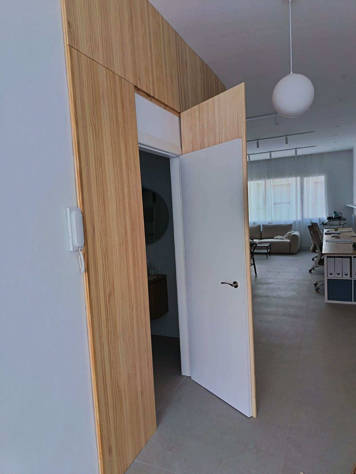 Una pared revestida de madera clara rodea una puerta blanca entreabierta que da acceso a una habitación con muebles de oficina y una lámpara de globo.