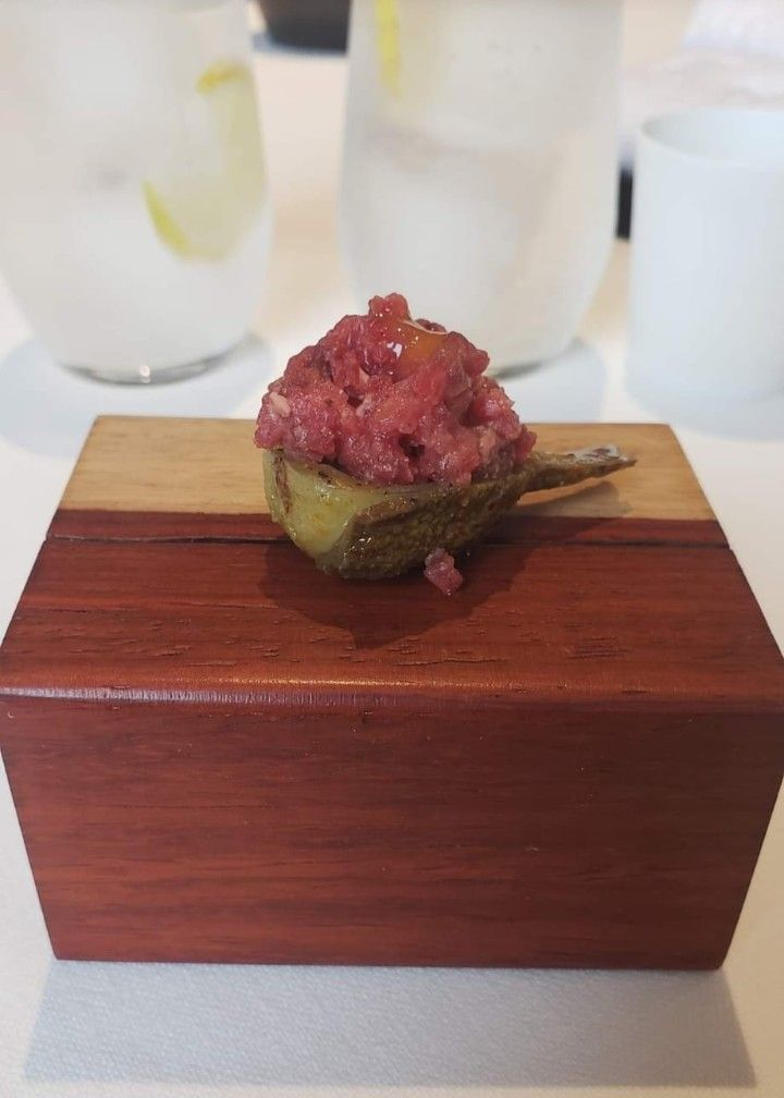 Un pequeño trozo de tartar de ternera servido sobre una verdura asada dentro de un bloque de madera, con vasos borrosos al fondo.