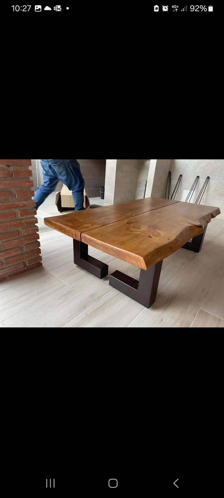 Una mesa de centro rústica de madera con borde natural y patas oscuras en forma de U, situada en una habitación con una pared de ladrillo visto de fondo.