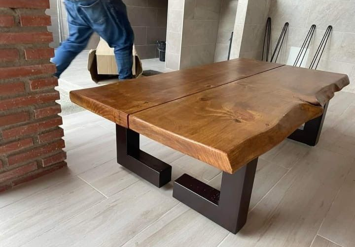 Una mesa de centro de madera con bordes naturales y patas de metal oscuro se alza sobre un suelo de baldosas en una habitación con una pared decorativa de ladrillo.