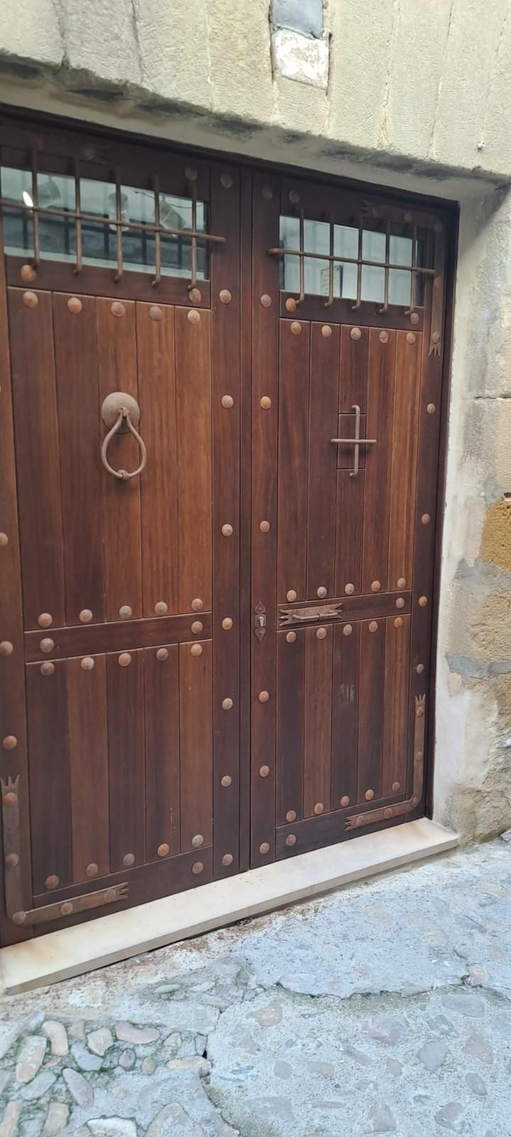 Una rústica puerta doble de madera con remaches de hierro, aldabas decorativas de metal y pequeñas ventanas, integrada en un muro de piedra.