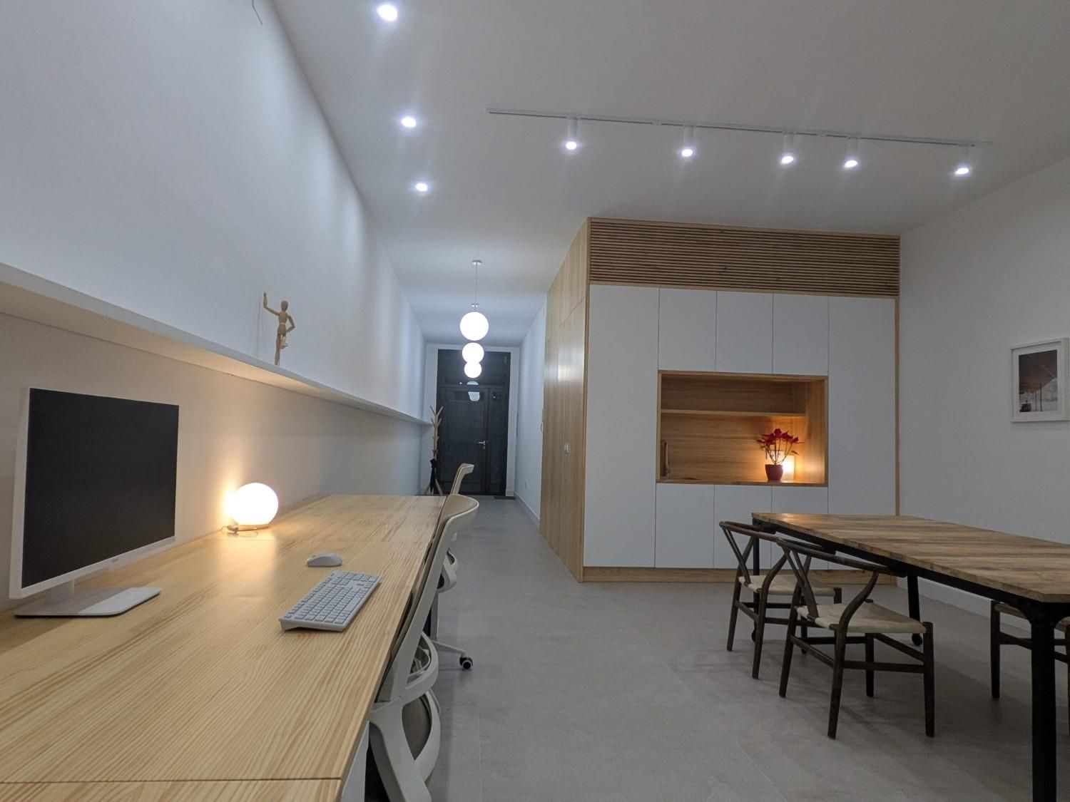 Un espacio de oficina moderno y minimalista con un escritorio largo de madera, una mesa de comedor, paredes blancas y un mueble de pared revestido de madera.