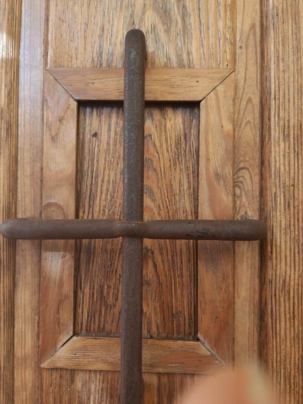 Un objeto metálico de color óxido, con forma de cruz, montado verticalmente sobre el panel de madera empotrado de una puerta.