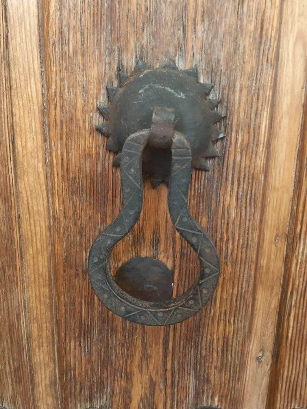 Un aldabón rústico de metal oscuro con una placa trasera en forma de engranaje, montado sobre una puerta de madera con vetas verticales.