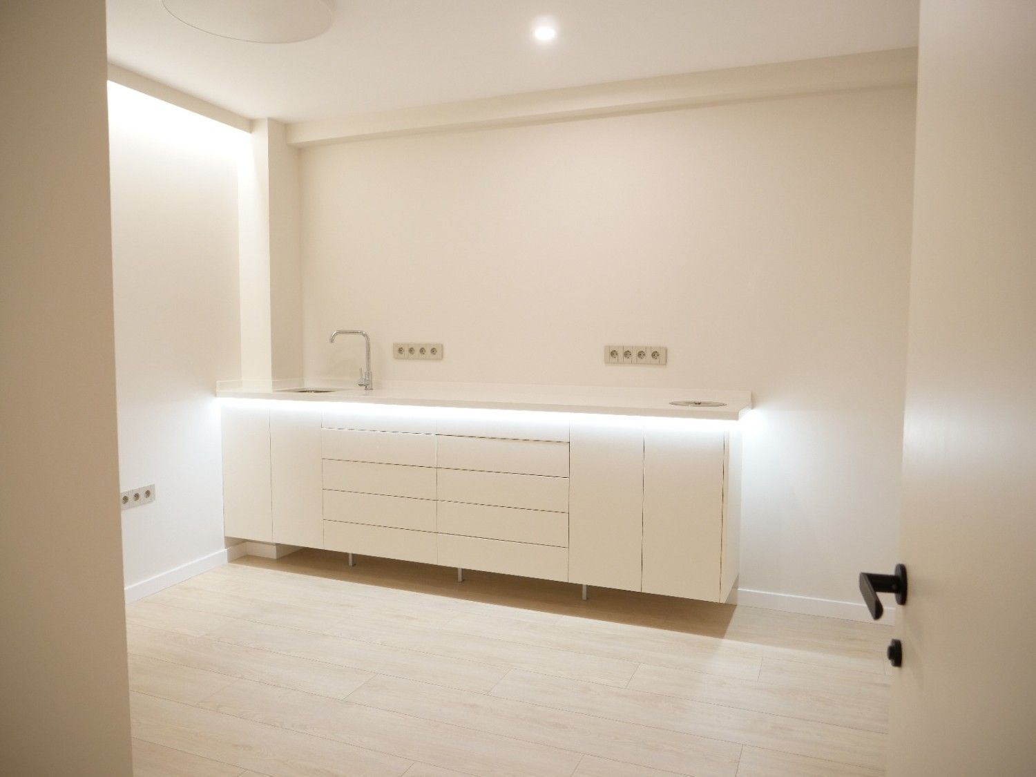 Una habitación blanca minimalista con una pequeña cocina empotrada en la pared, iluminación LED mediante tiras y un grifo cromado individual.