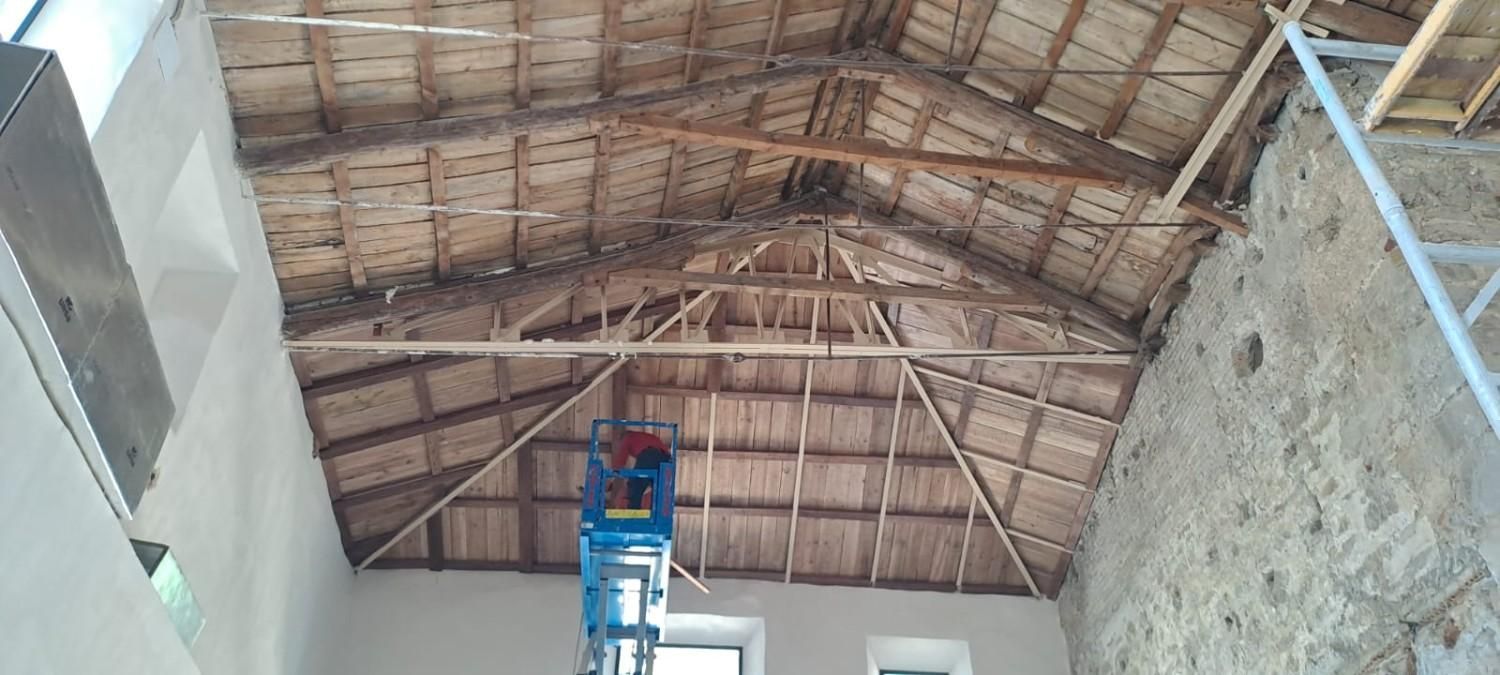 Una plataforma elevadora de tijera azul se extiende hacia una estructura de techo de madera en un edificio en renovación.
