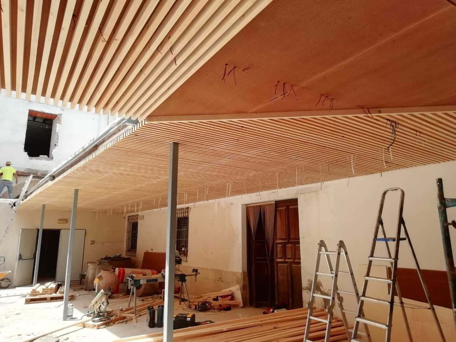 Los obreros de la construcción instalan un techo de listones de madera bajo un patio exterior utilizando escaleras y materiales de construcción.