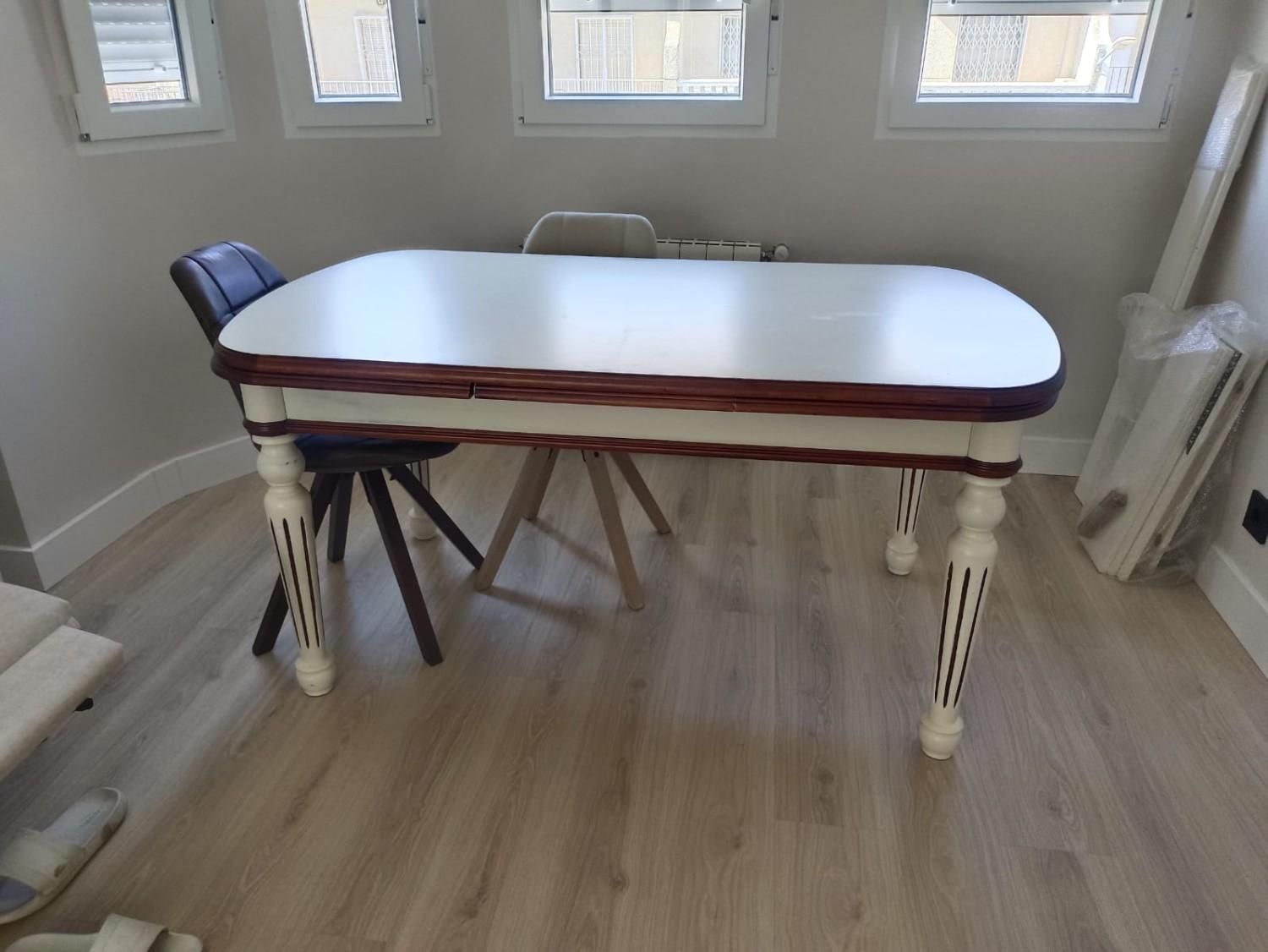 Sobre un suelo de madera clara, se alza una mesa rectangular con tablero blanco y detalles de madera oscura, con patas blancas ornamentadas.