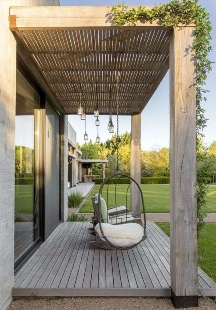 Una terraza de madera con techo de pérgola de listones, con una silla colgante en forma de huevo, con vistas a un jardín verde al atardecer.