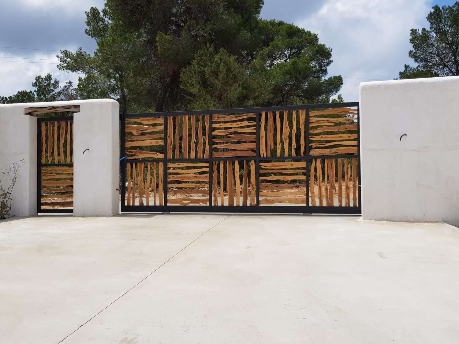 Una puerta corredera de acceso para vehículos y una puerta peatonal adyacente con marcos de metal negro y listones de madera verticales entre paredes blancas.