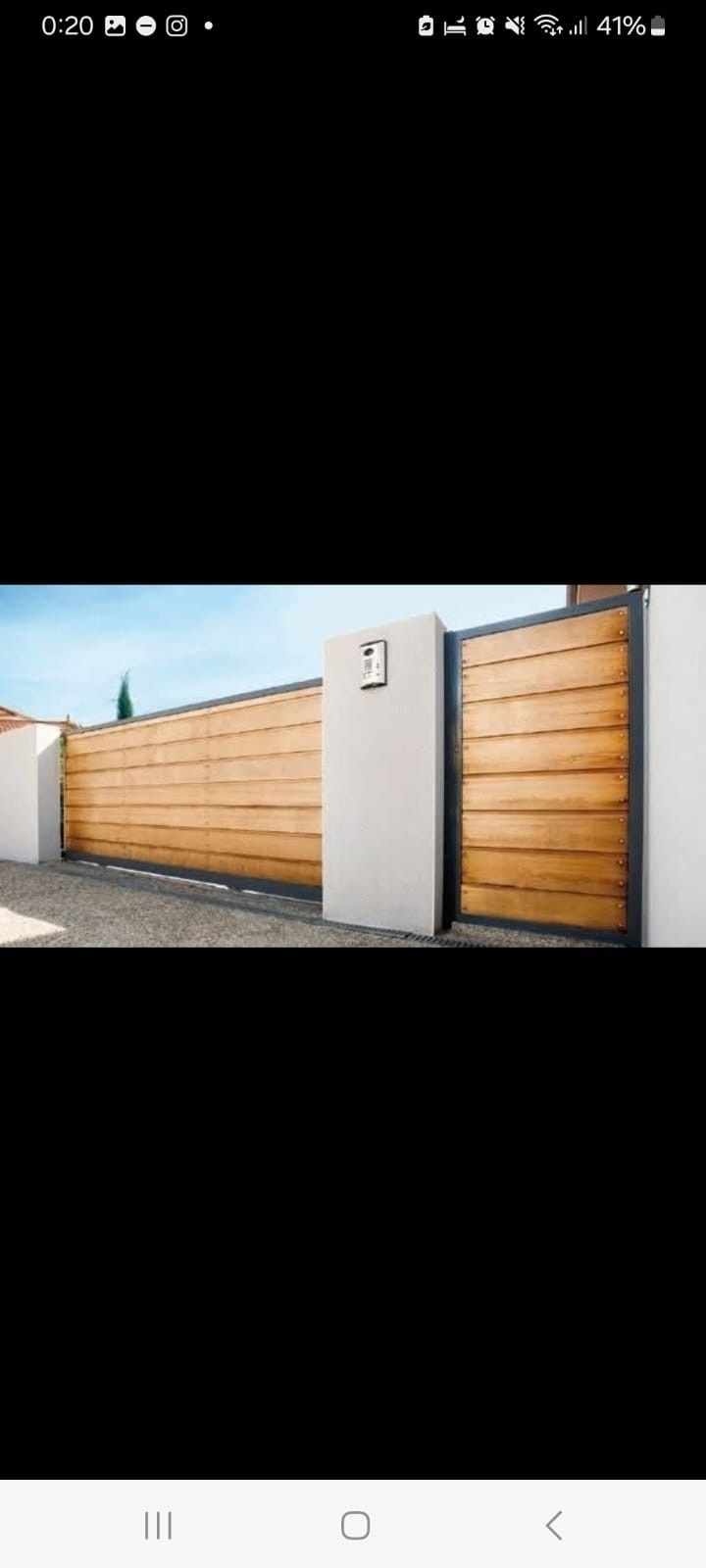 Moderna puerta de entrada para vehículos con tablones horizontales de madera clara enmarcados en acero negro, situada entre pilares de piedra blanca.