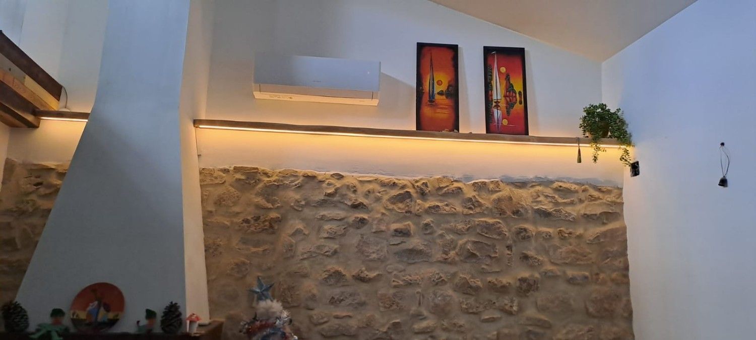 Un muro de piedra con una repisa iluminada que sostiene dos cuadros enmarcados y una pequeña planta, bajo un techo abovedado blanco.