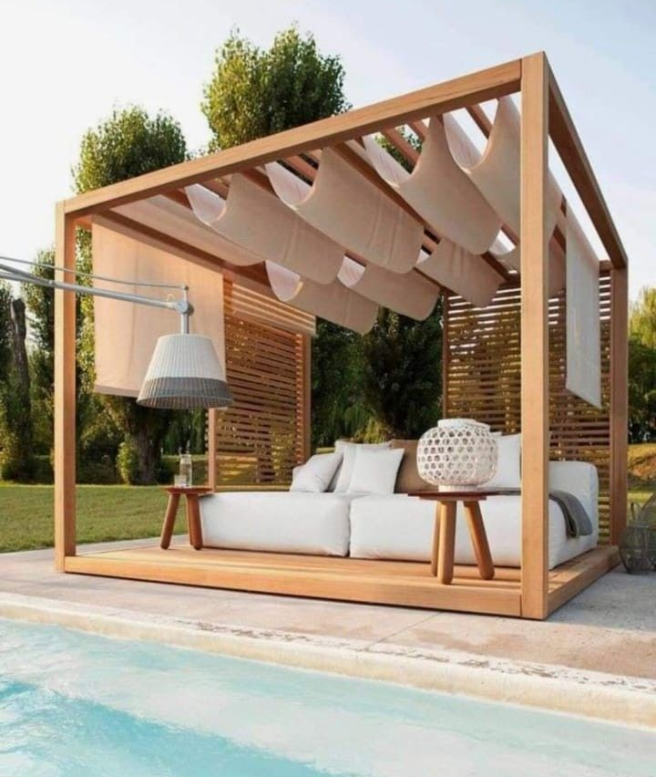 Una moderna cabaña de madera con cojines blancos, un techo de tela drapeada y un entorno junto a la piscina al atardecer.