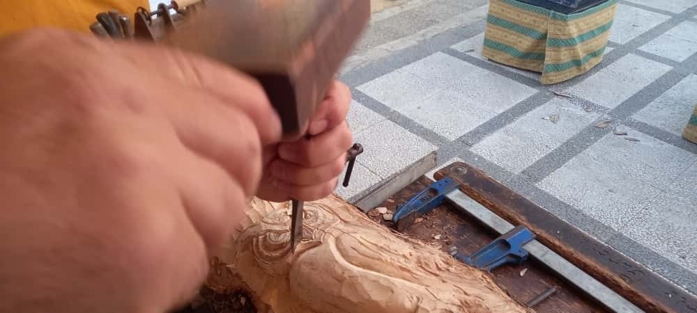 Una persona utiliza un mazo de madera para golpear un cincel de metal contra un trozo de madera sujeto con abrazaderas en un banco de trabajo.