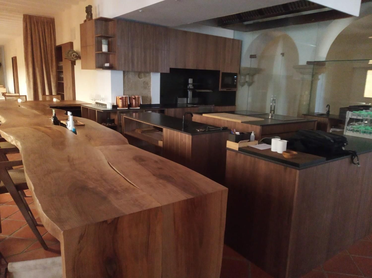 Una cocina moderna cuenta con una larga barra de madera con borde natural en primer plano, armarios de madera oscura y puertas arqueadas.