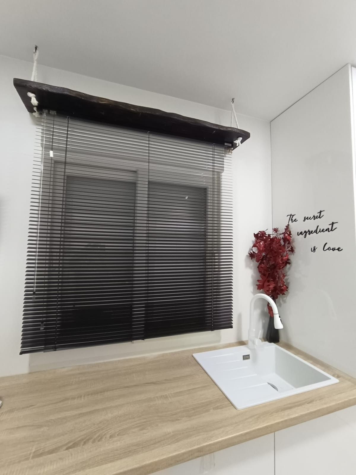 Un fregadero de cocina con grifo blanco se encuentra debajo de una ventana con persianas horizontales negras, adornada con un ramo de flores rojas.