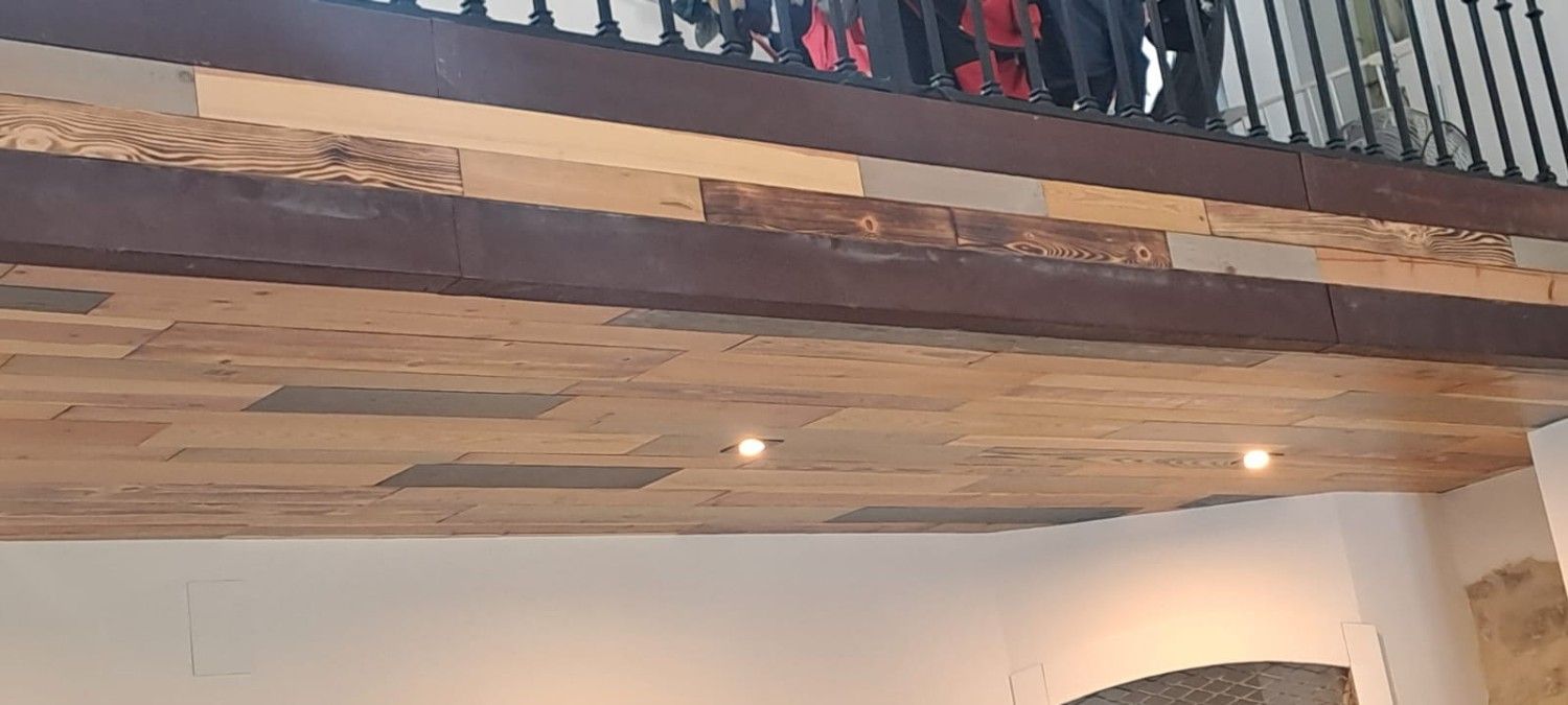 Un techo de madera hecho de tablones con dos luces empotradas, debajo de un balcón con barandillas de metal oscuro.