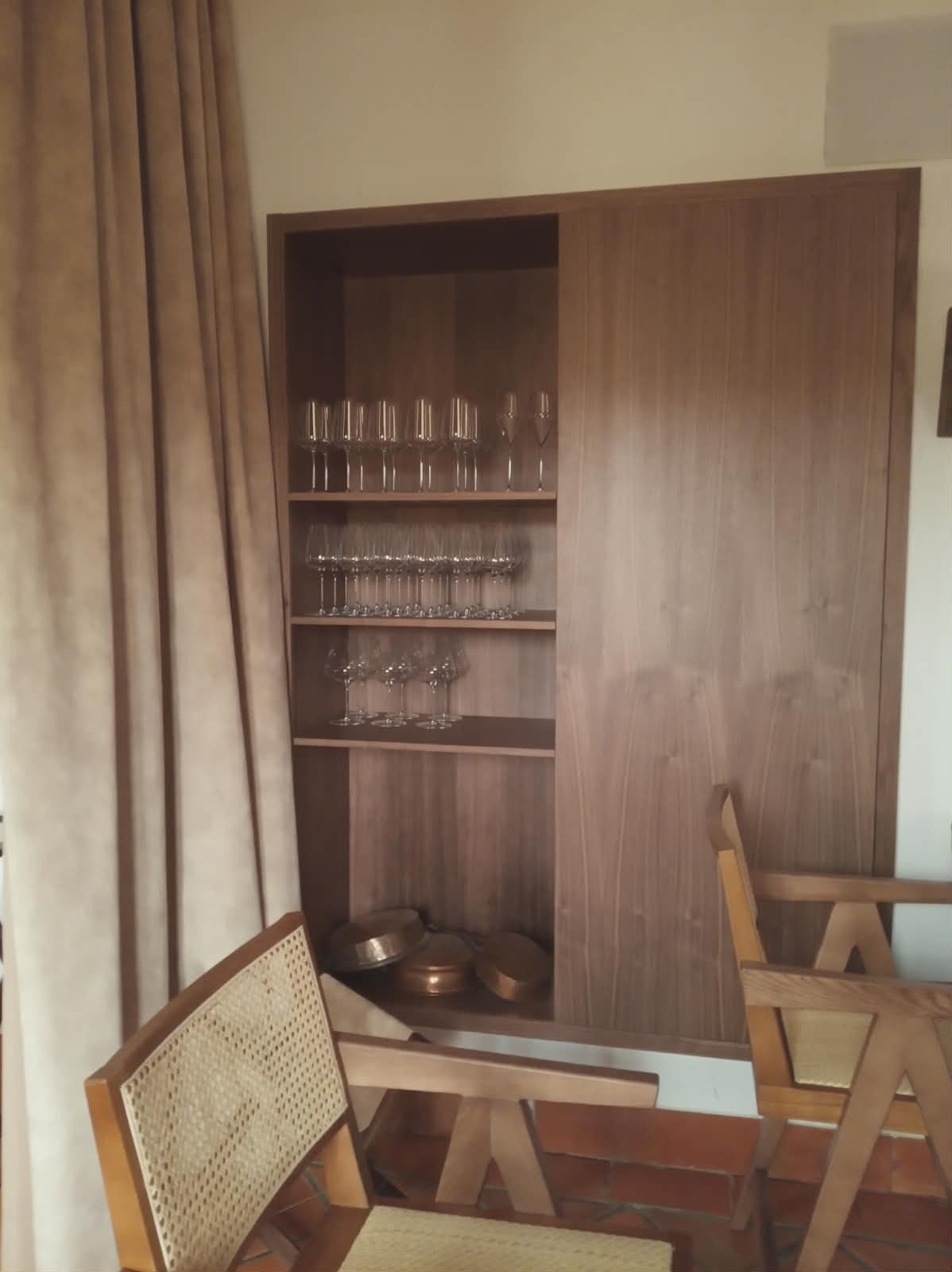 Un armario de madera con estantes abiertos que exhiben filas de cristalería, situado junto a una cortina y dos sillas de madera.