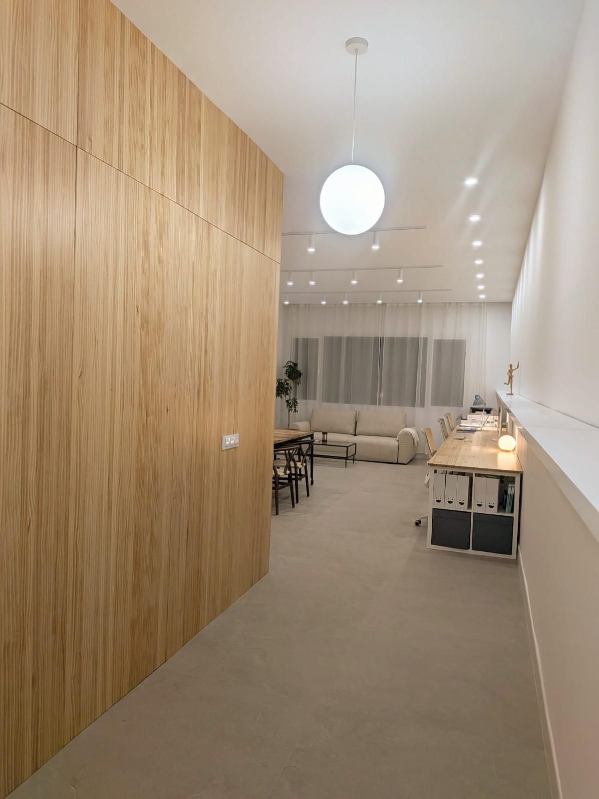 Un largo pasillo conduce a una sala de estar con una pared revestida de paneles de madera clara a la izquierda, paredes blancas y una moderna lámpara de globo.