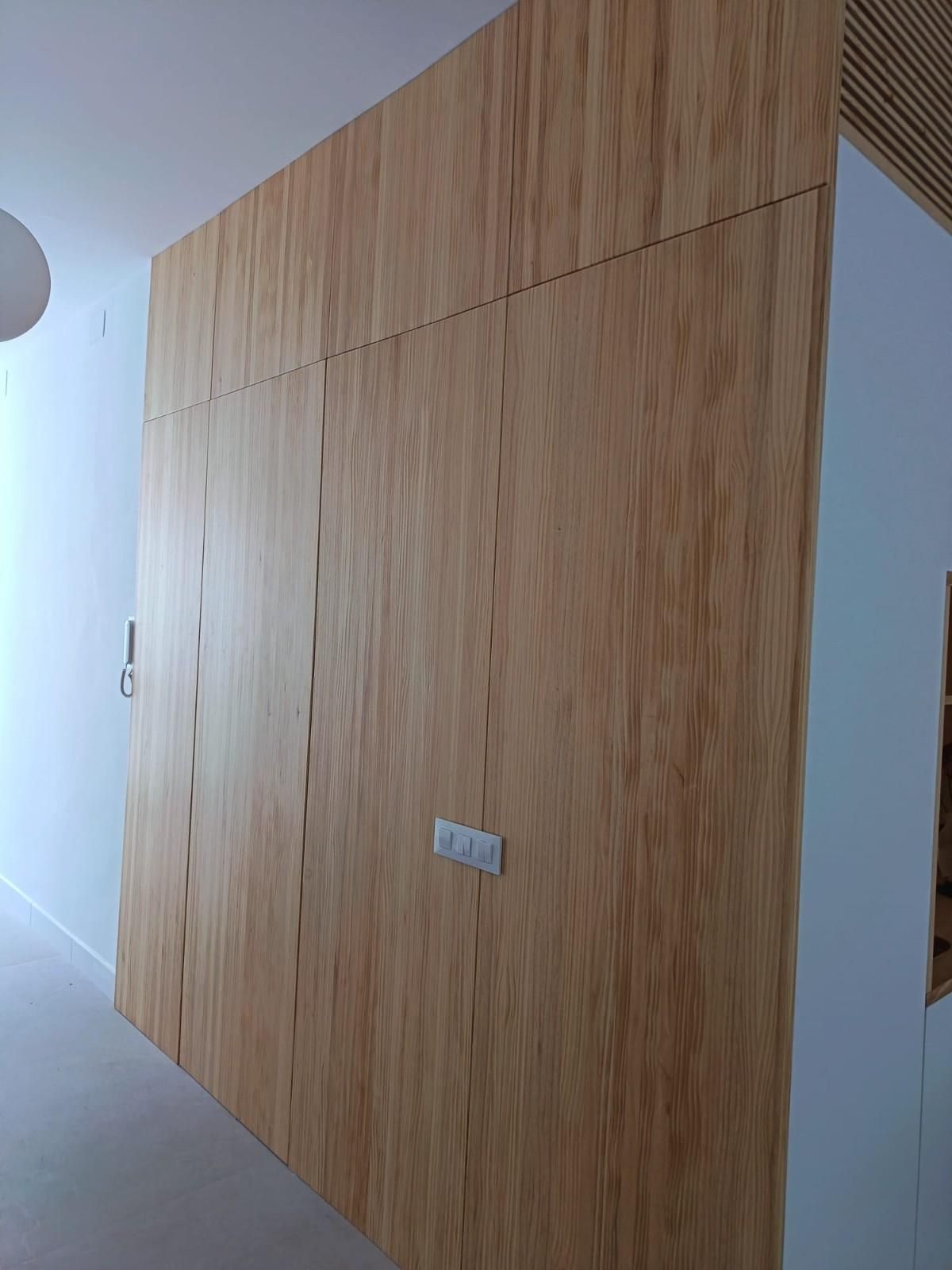 Una sección de pared revestida con paneles verticales de madera de roble claro que incorpora un interruptor de luz.