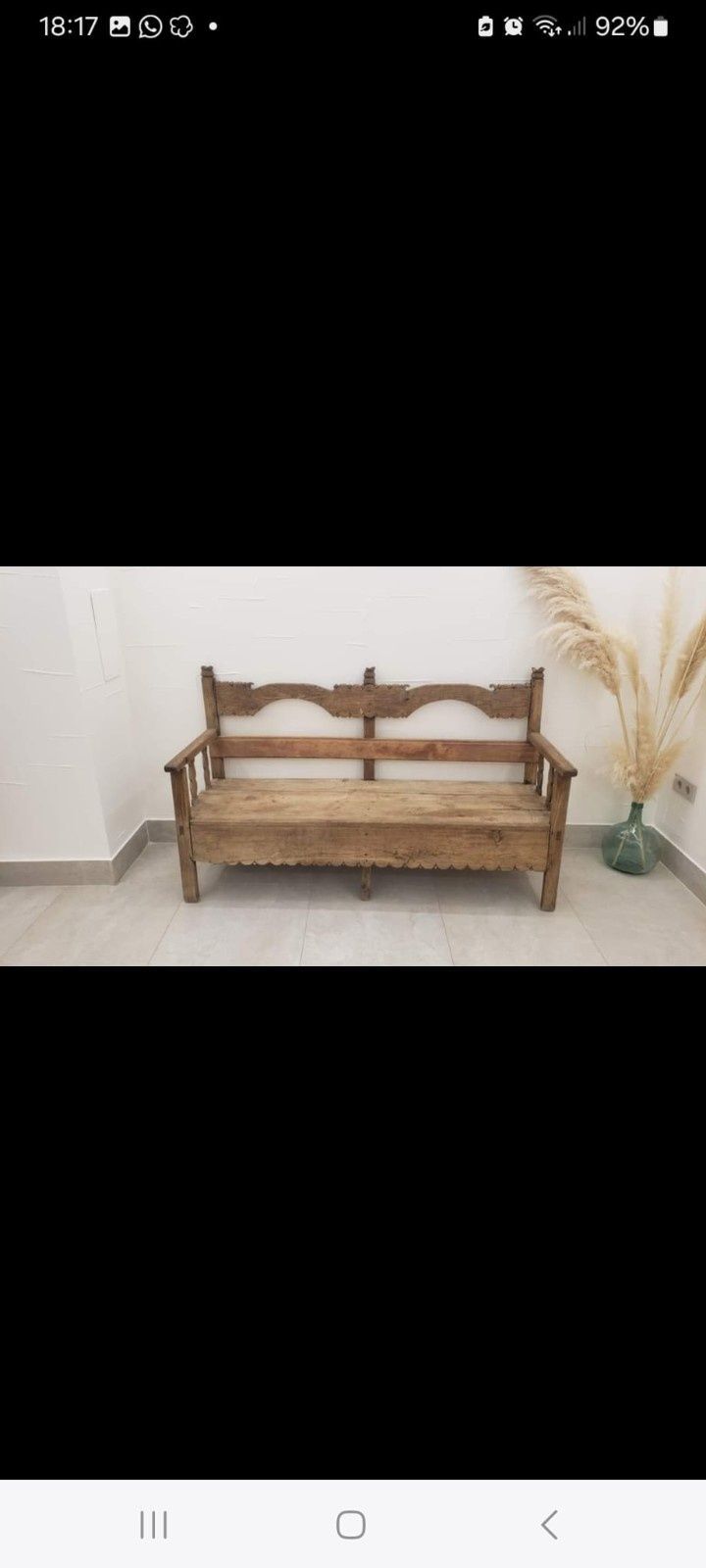 Un banco rústico de madera tallada con un respaldo alto se alza contra una pared blanca lisa adornada con un arreglo floral de flores secas.