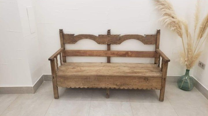 Un banco rústico de madera con detalles festoneados se alza contra una pared blanca, junto a un jarrón con hierba de la pampa seca.