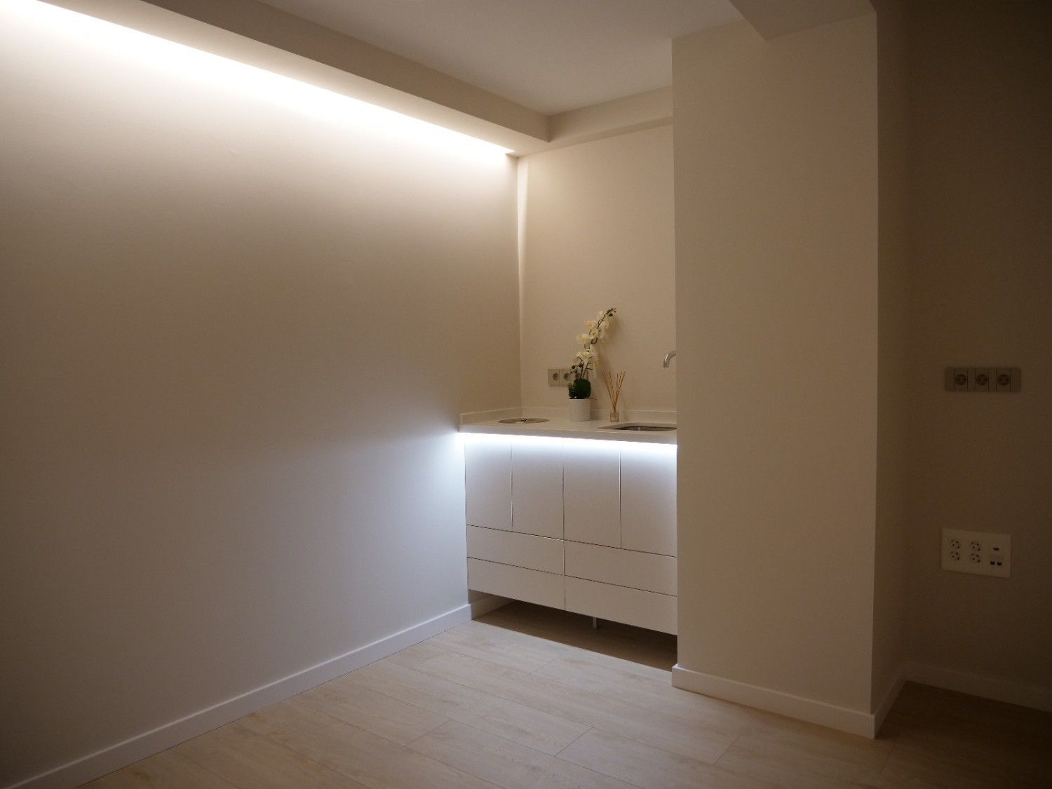 Una habitación moderna y minimalista con un mueble de tocador blanco empotrado que cuenta con iluminación cálida debajo del mueble y luces empotradas en la pared.
