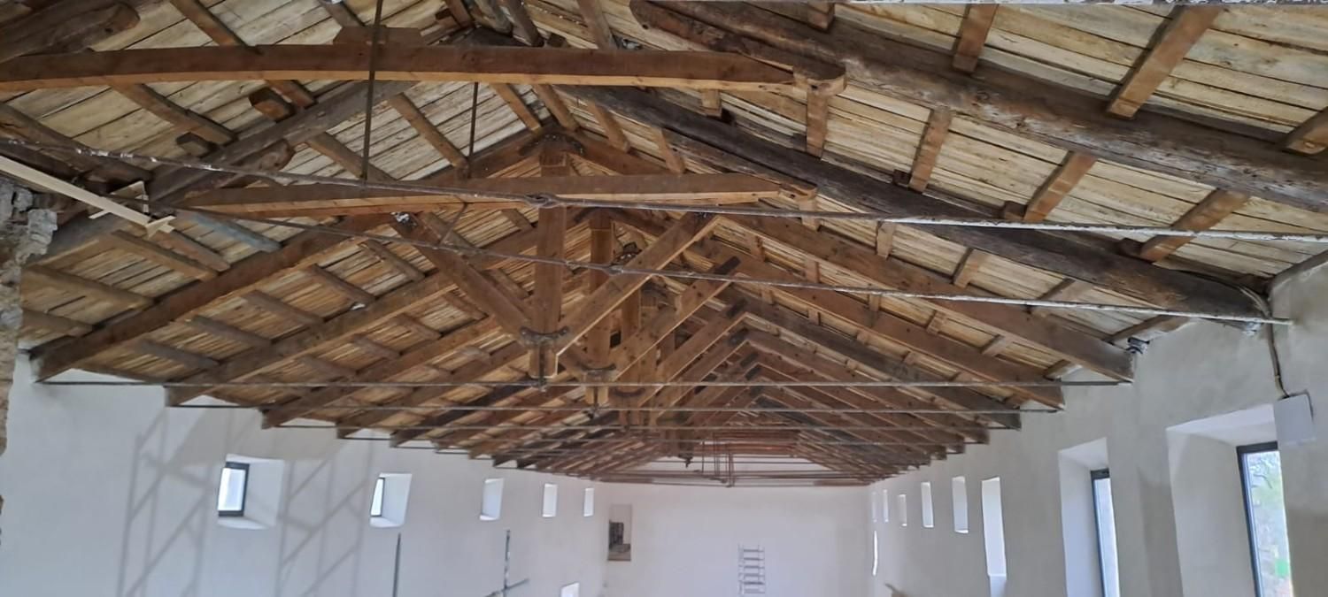 El interior de un edificio rústico con paredes blancas y un techo de vigas de madera a la vista, con pequeñas ventanas espaciadas uniformemente.