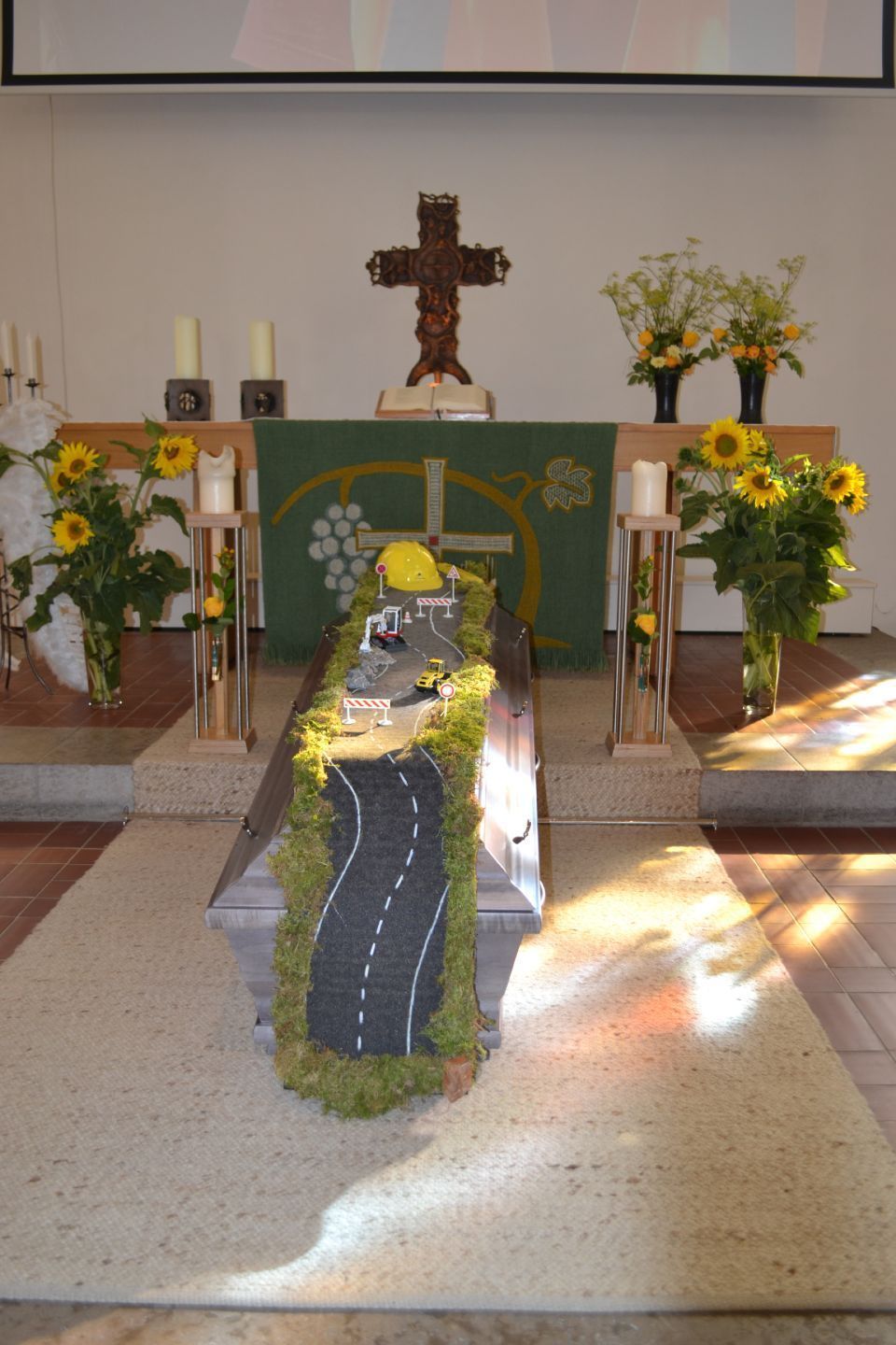 Altar mit dem Sarg dekoriert mit Sonnenblumen, Kerzen und einer Straße über dem geschlossenen Sarg mit Bauarbeiterhelm, Moos und Mini-Baustelle.