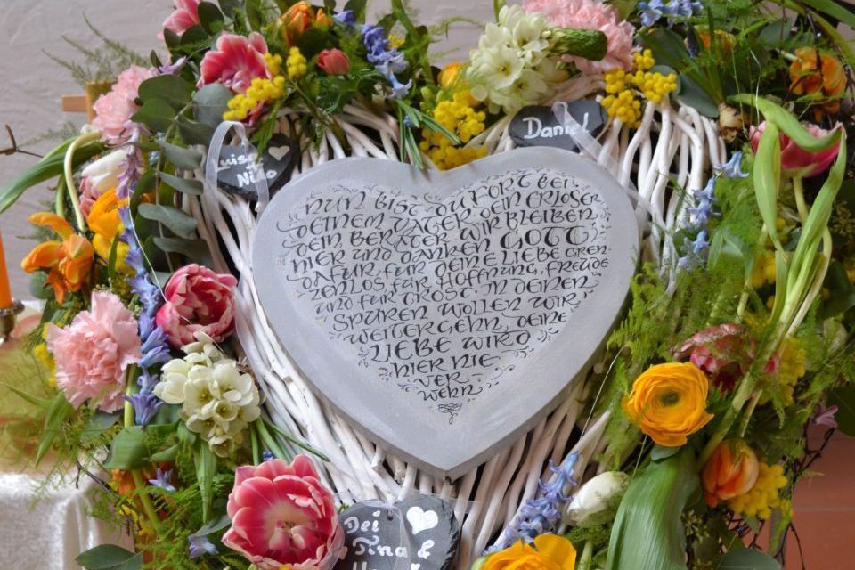 Blumengesteck in Form eines Herzen aus Ästen und Wildblumen, mit einem beschrifteten Herz in der Mitte.