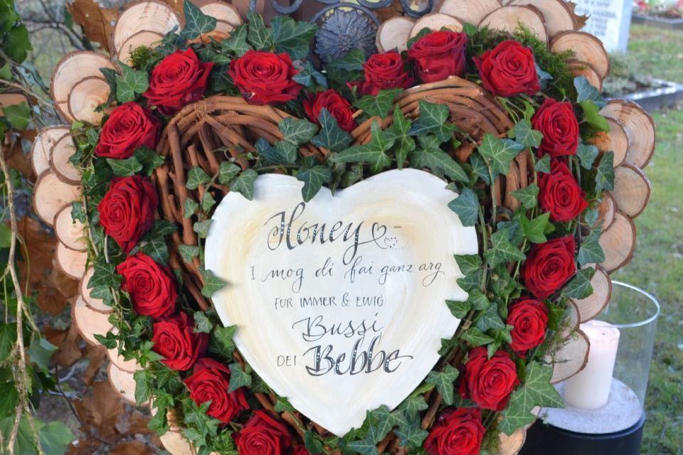 Aufgestelltes Blumengesteck in Herzform mit Rosen, Efeu, Holz und einem Spruch.